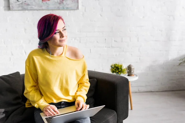 Hipster girl with colorful hair sitting on sofa, using computer, smiling and looking away — Stock Photo