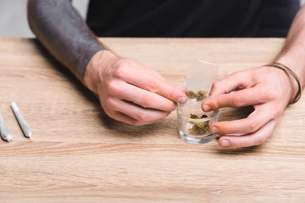 Cropped view of young man rolling joint with weed while sitting behind table — Stock Photo