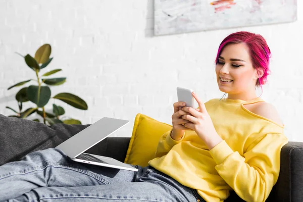 Hipster menina em roupas casuais sentado no sofá, sorrindo e usando smartphone — Fotografia de Stock