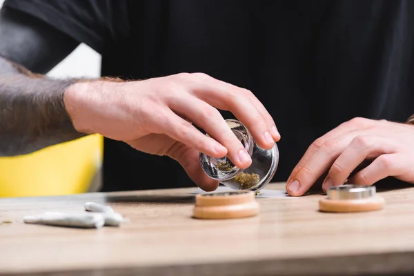 Vista recortada del hombre poniendo cannabis medicinal en el papel de tabaco mientras está sentado en la mesa con las articulaciones - foto de stock