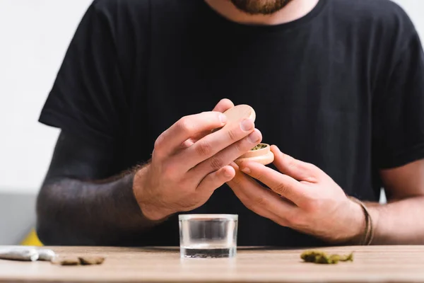 Visão cortada do homem que fecha o moedor de ervas com maconha medicinal enquanto está sentado à mesa — Fotografia de Stock