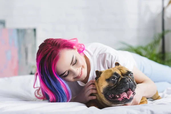 Menina bonita com cabelo colorido deitado na cama, sorrindo e acariciando bulldog francês — Fotografia de Stock