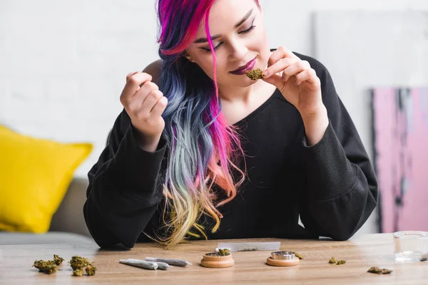 Fille hipster avec des cheveux colorés reniflant la marijuana médicale et assis derrière la table avec des articulations et broyeur d'herbes — Photo de stock