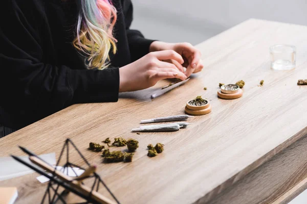 Vista cortada da mulher rolando conjunta enquanto sentado à mesa com cannabis medicinal, moedor de ervas e articulações — Fotografia de Stock