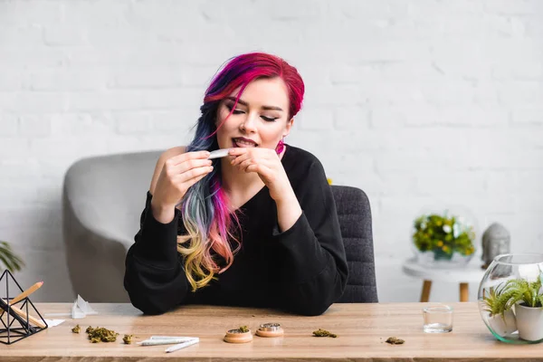 Attractive girl licking joint with medical marijuana while sitting behind table in living room — Stock Photo