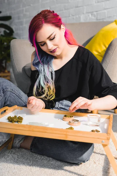 Belle fille avec des cheveux colorés assis sur le sol derrière une petite table et faisant joint avec de l'herbe — Photo de stock