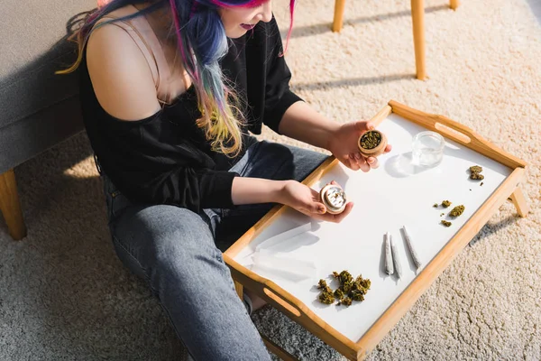 Vista cortada da menina com cabelos coloridos sentados no chão atrás de pequena mesa com moedor de ervas nas mãos na sala de estar — Fotografia de Stock