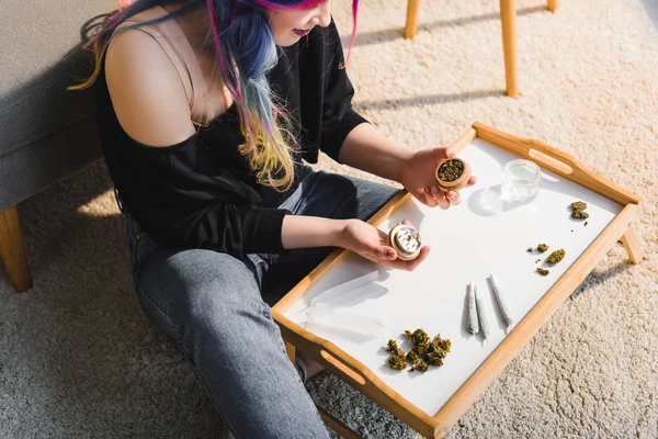 Cropped view of girl with colorful hair sitting on floor behind small table with herb grinder in hands in living room — Stock Photo