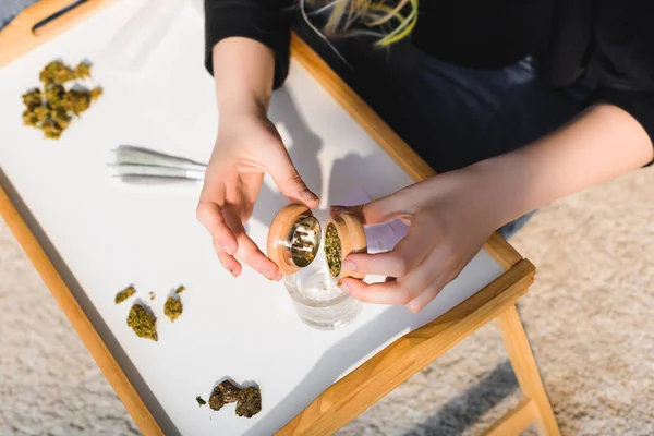 Top view of girl putting weed from marijuana grinder in glass on wooden tray — Stock Photo