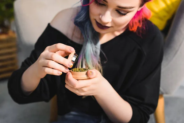 Beautiful girl with colorful hair holding and looking at herb grinder while sitting on floor — Stock Photo