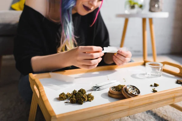 Enfoque selectivo de la niña rodando conjunta y marihuana medicinal, articulaciones y amoladora de hierbas en la mesa - foto de stock