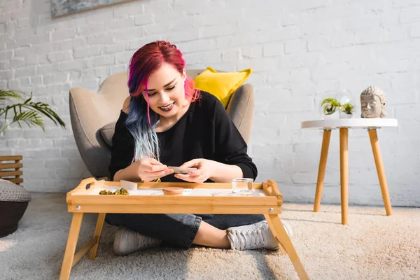 Attractive hipster girl sitting on floor behind table and rolling joint — Stock Photo