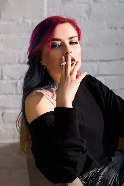 Hermosa chica con el pelo colorido sentado en el sillón, fumar y mirando a la cámara - foto de stock