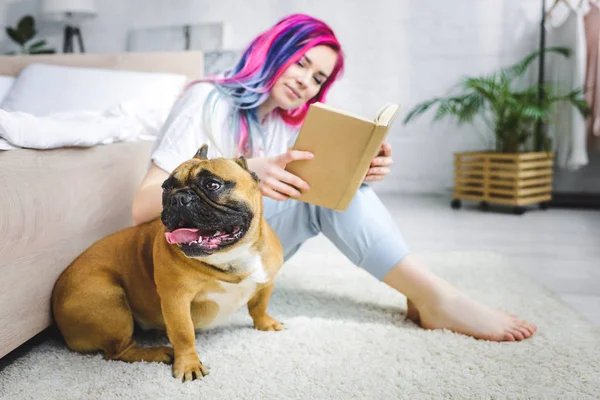 Beautiful girl with colorful hair reading book and sitting on floor near French bulldog — Stock Photo