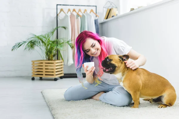 Chica atractiva con pelo colorido sosteniendo taza de café, sentado en el suelo y acariciando bulldog lindo - foto de stock