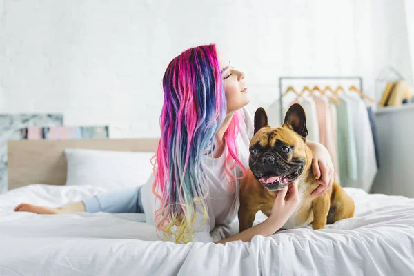 Chica con colorido pelo acariciando lindo bulldog francés y mirando hacia otro lado mientras está acostado en la cama - foto de stock
