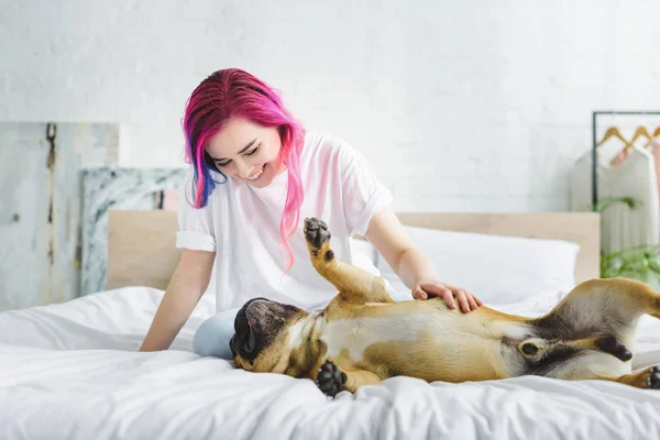 Girl with colorful hair petting and looking at cute french bulldog laying on back in bed — Stock Photo