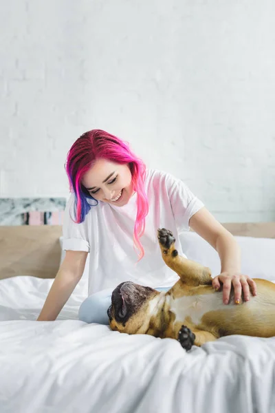 Menina com cabelo colorido acariciando e olhando para buldogue francês bonito, que deitado de volta na cama — Fotografia de Stock