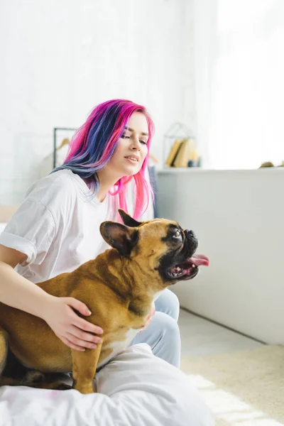 Mädchen mit buntem Haar streichelt Bulldogge und schaut weg, während sie auf dem Bett sitzt — Stockfoto