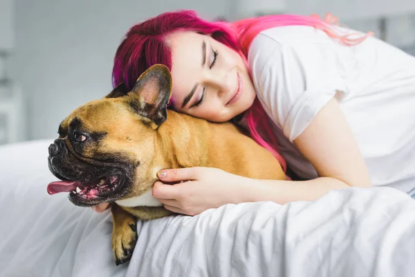 Красивая девушка с красочными волосами, обнимающая милый бульдог, лежа в постели — стоковое фото