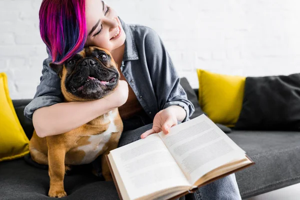 Chica con pelo colorido abrazando bulldog, sosteniendo libro y sentado en el sofá - foto de stock