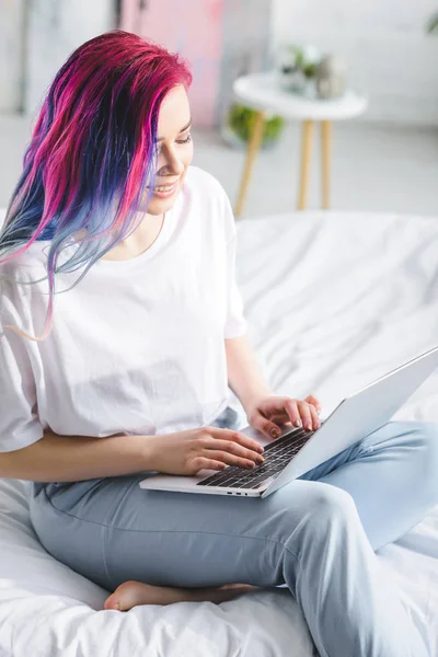 Chica atractiva con el pelo colorido sentado en la cama, sonriendo y utilizando el ordenador portátil - foto de stock