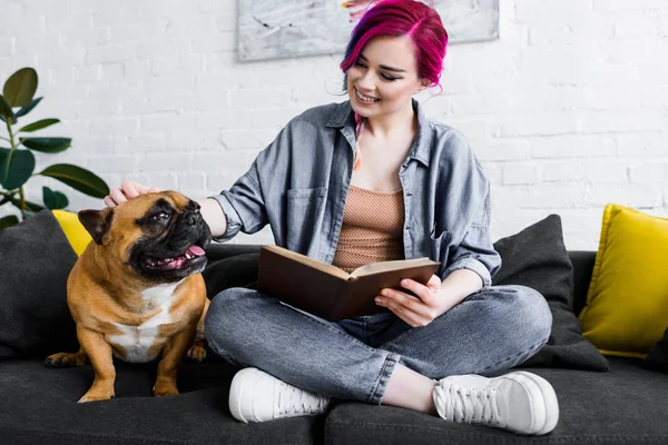 Menina bonita com cabelo colorido sentado no sofá com livro e acariciando bulldog bonito — Fotografia de Stock