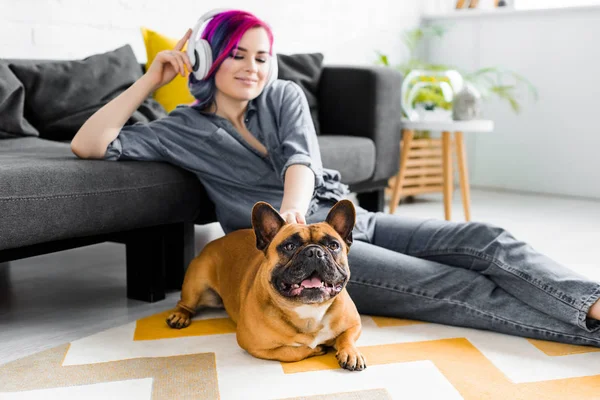 Beautiful girl with colorful hair and headphones sitting on floor and petting french bulldog — Stock Photo