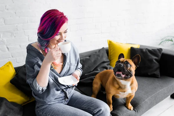Beautiful girl with colorful hair drinking coffee and sitting on sofa near cute french bulldog — Stock Photo