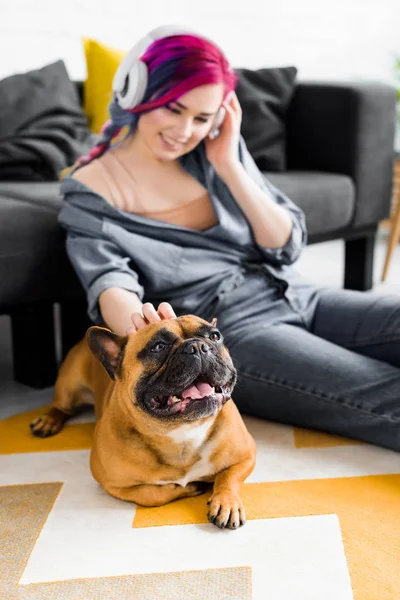 Foco seletivo da menina bonita com cabelos coloridos e fones de ouvido sentado no chão e acariciando bulldog francês — Fotografia de Stock
