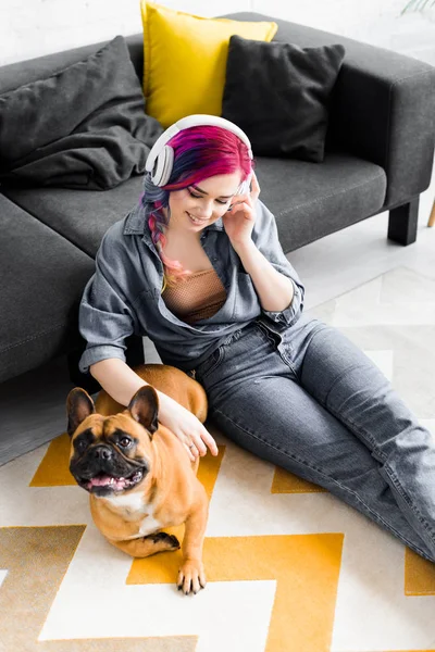 Vista de ángulo alto de hermosa chica con el pelo colorido y auriculares sentados en el suelo y acariciar bulldog francés - foto de stock