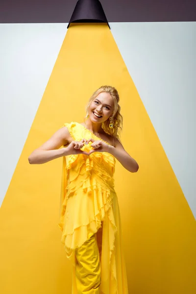 Щаслива молода жінка дивиться на камеру і показує серце руками на білому і жовтому — стокове фото