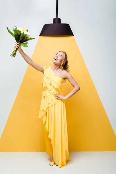 Attractive young woman standing with hand on hip and holding bouquet of flowers on white and yellow — Stock Photo