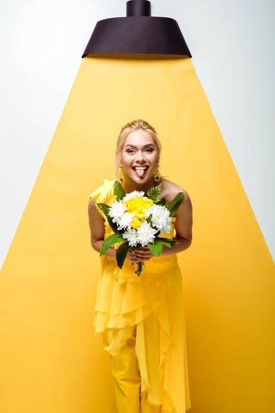 Glückliche junge Frau zeigt Zunge und hält Blumenstrauß auf weiß und gelb — Stockfoto