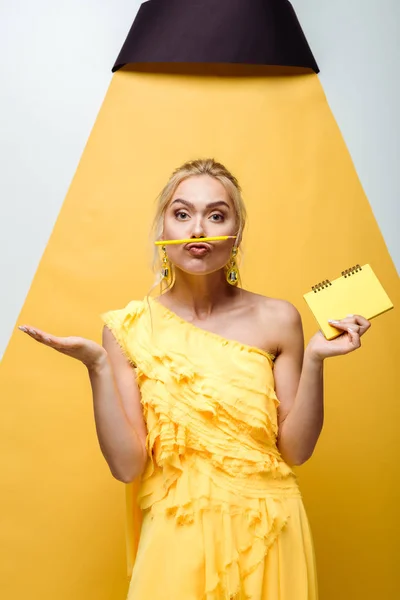 Jeune femme blonde avec le visage de canard tenant crayon près de la bouche et carnet à la main sur blanc et jaune — Photo de stock