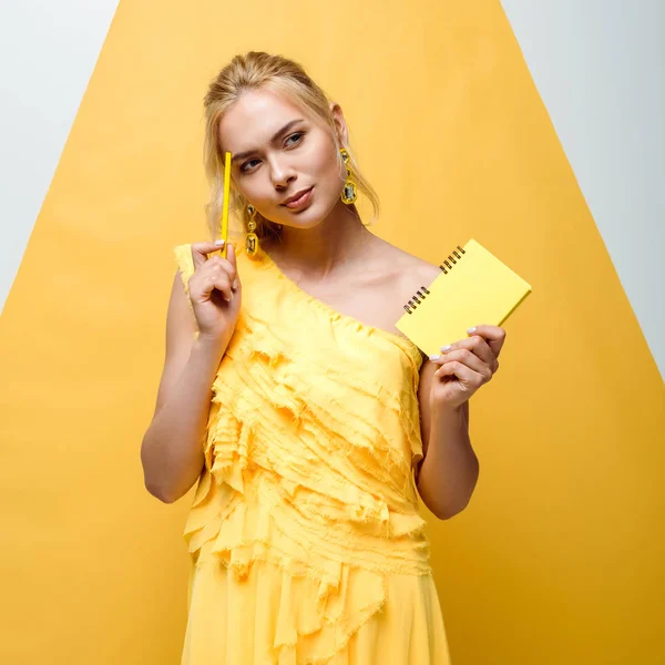 Niña rubia reflexiva sosteniendo cuaderno y lápiz en blanco y amarillo - foto de stock