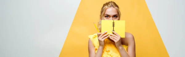 Panoramic shot of blonde young woman covering face with notebook on white and yellow — Stock Photo