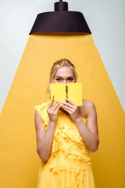 Blonde young woman covering face with notebook on white and yellow — Stock Photo