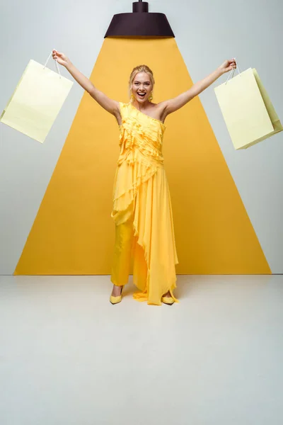Allegra donna bionda che tiene borse della spesa mentre posa su bianco e giallo — Foto stock