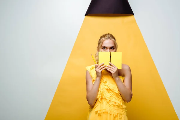 Young blonde woman covering face with notebook on white and yellow — Stock Photo