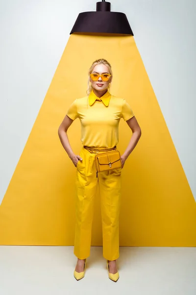 Jolie femme blonde en lunettes de soleil debout avec les mains dans les poches sur blanc et jaune — Photo de stock