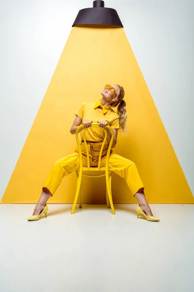 Femme blonde gaie dans des lunettes de soleil assis sur une chaise et regardant la lampe sur blanc et jaune — Photo de stock