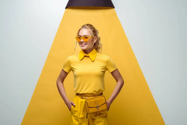 Fröhliche blonde Frau mit Sonnenbrille, die Hände in den Taschen auf weiß und gelb — Stockfoto