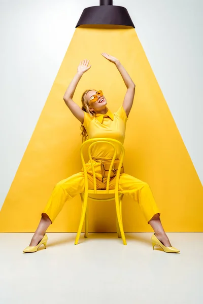 Happy blonde woman in sunglasses sitting on chair with hands above head and looking at lamp on yellow and white — Stock Photo