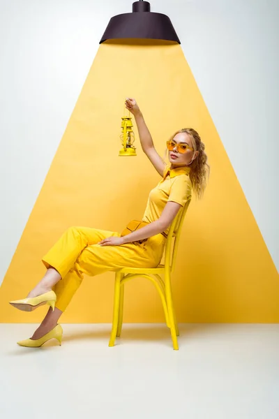 Attractive blonde woman in sunglasses sitting on chair and holding vintage lamp on white and yellow — Stock Photo