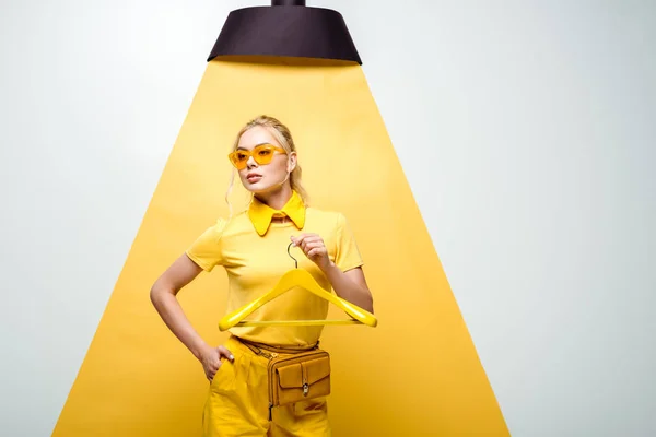 Attractive woman in sunglasses holding hanger and standing with hand in pocket on white and yellow — Stock Photo