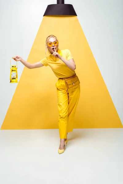Donna bionda in occhiali da sole posa e mostra un segno di silenzio mentre tiene in mano la lampada vintage su bianco e giallo — Foto stock