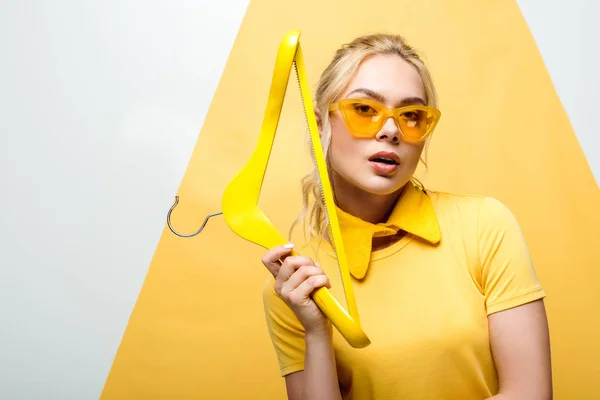 Jolie femme blonde en lunettes de soleil tenant cintre en bois sur blanc et jaune — Photo de stock