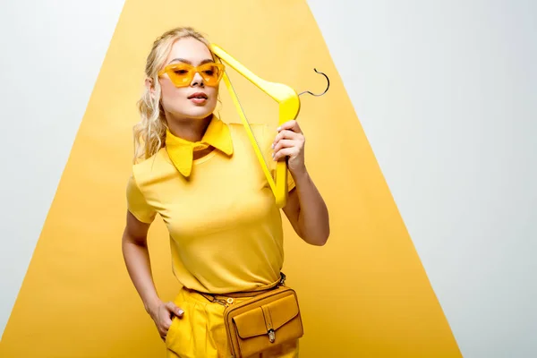 Attractive blonde woman in sunglasses posing with hand in pocket and holding hanger on white and yellow — Stock Photo