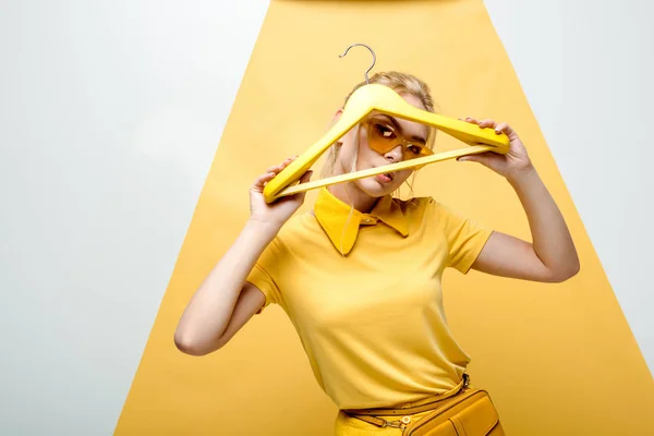 Attractive blonde woman in sunglasses covering face while holding hanger on white and yellow — Stock Photo